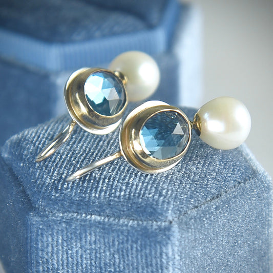 Pair of earrings with blue gemstones and pearls on a blue cushioned stand.