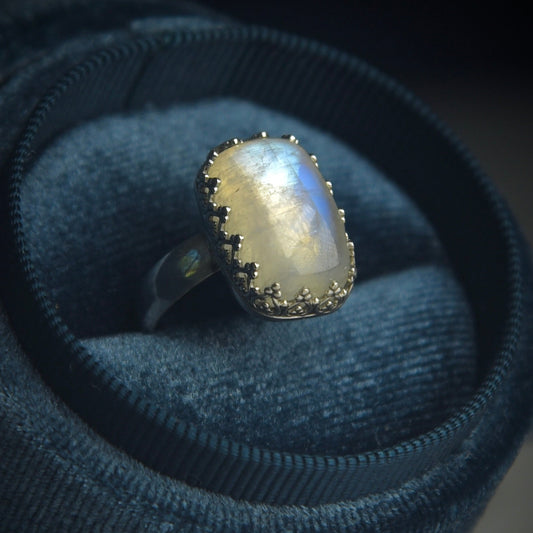 A silver ring with a large, oval-shaped iridescent moonstone cabochon, displayed in a dark blue velvet ring roll against a dark background.