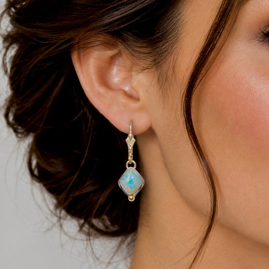 A close-up of a model's ear wearing a gold drop earring with a light blue opal gemstone.