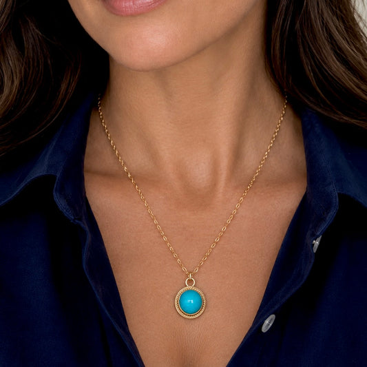 A close-up of a woman wearing a delicate gold chain necklace with a round turquoise pendant, styled with a dark navy blue collared shirt.
