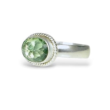 A silver ring featuring an oval-cut light green gemstone in a textured bezel setting, shown on a plain white background.