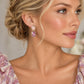 A close-up of a woman wearing a pair of dangling earrings with oval pink gemstones in gold-toned settings, shown against a softly blurred neutral background.