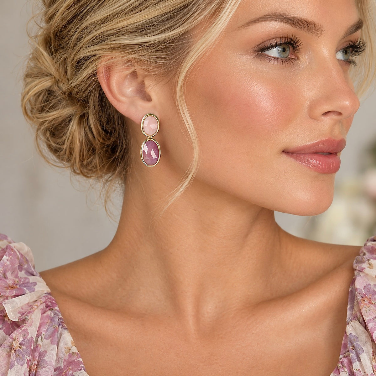 A close-up of a woman wearing a pair of dangling earrings with oval pink gemstones in gold-toned settings, shown against a softly blurred neutral background.