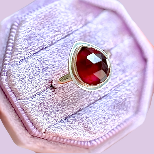 A gold ring with a faceted deep red gemstone, displayed inside a light pink velvet ring box.