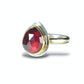 A teardrop-shaped faceted red gemstone ring set in a gold-toned bezel on a polished silver band, shown on a white background with a soft shadow.